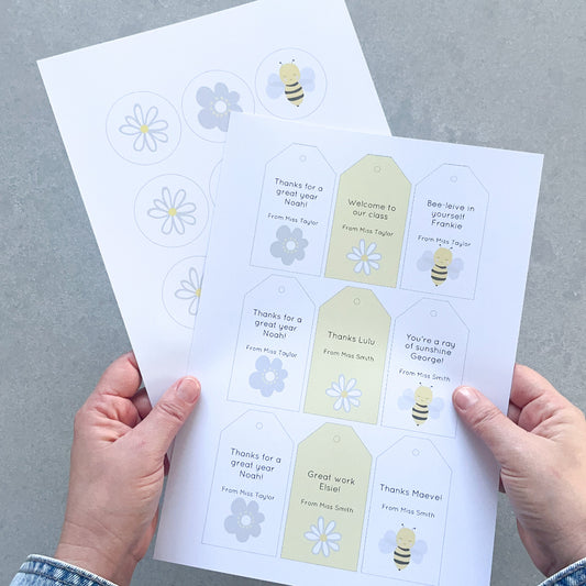 Sheet of printed name tags with bee and flower designs held by hands on a grey background