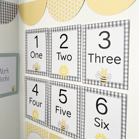 Educational number posters with bee and sunflower graphics on a checkered background displayed on a white wall.