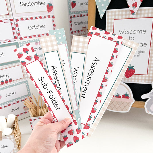 Hand holding educational classroom folder labels with strawberry design on a desk.
