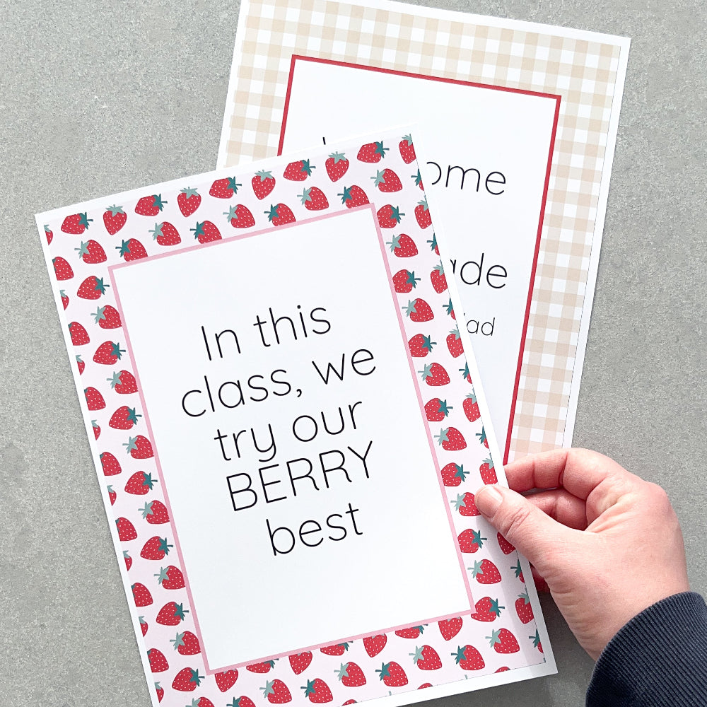 Two printed classroom posters with strawberry pattern and text on a grey surface, held by a hand.