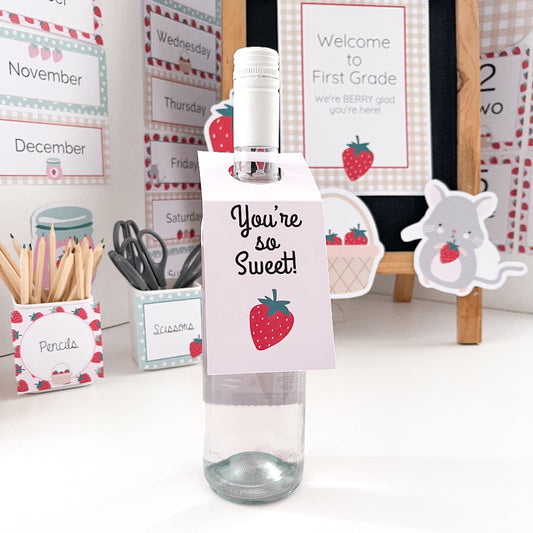 Bottle with a decorative tag with a strawberry on it. The. bottle surrounded by classroom supplies and decor.