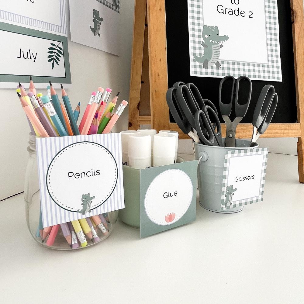 Square printable classroom labels in a crocodile theme on white bench with school supplies in caddies.