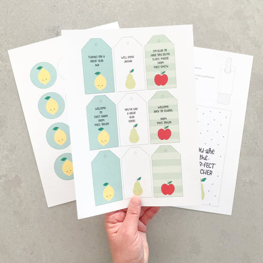 Hand holding a sheet of printed gift tags with lemon and apple designs on a gray surface.