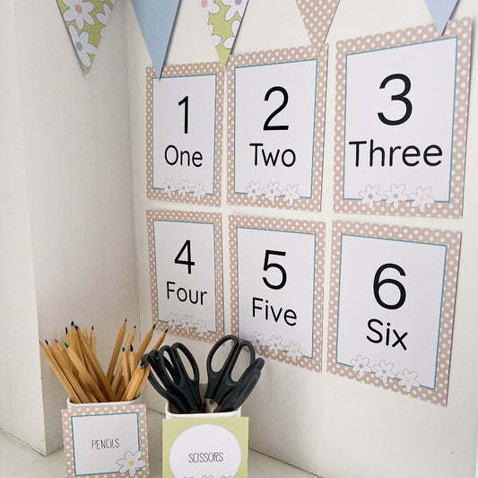 Children's educational wall chart with numbers 1-6. In the foreground there is caddies labeled 'Pencils' and 'Scissors' on a light background.