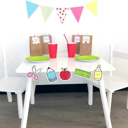 A set of party decorations featuring school-themed icons, including a white table with a green apple, pink and red cups, and decorative cutouts in the shape of a teacher's apple, scallop, triangle, and flag, with a 'Back to School' sign in the background.