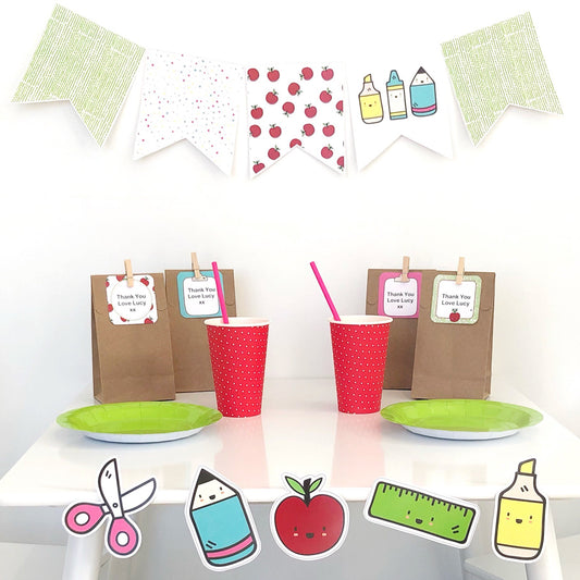 Brightly coloured school supply cut outs and bunting decoration on a white table. There is red cups, green plates and party bags with editable labels in the background.