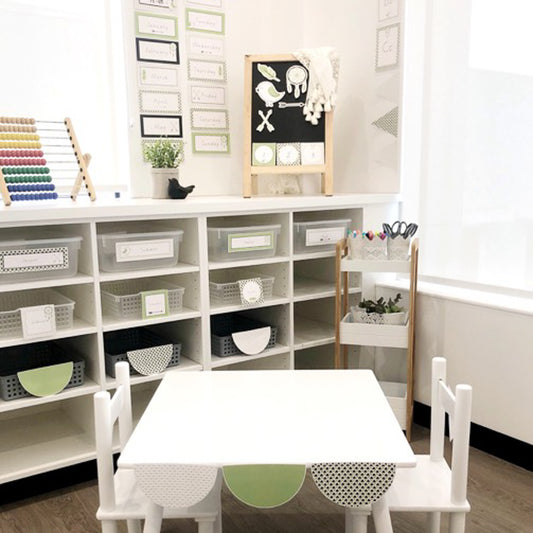 A classroom setting featuring a white table with green circle accents, surrounded by shelves with educational decor and supplies.