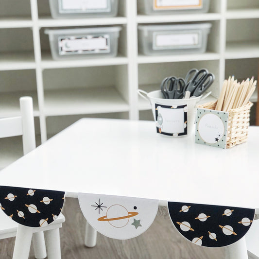 Space theme scalloped bunting displayed on the edge of a white table with classroom decor in the backgound.