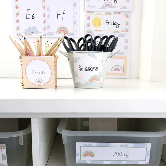 Classroom display with labels and charts in a pastel rainbow theme. There are caddies with scissors and pencils in the foreground.