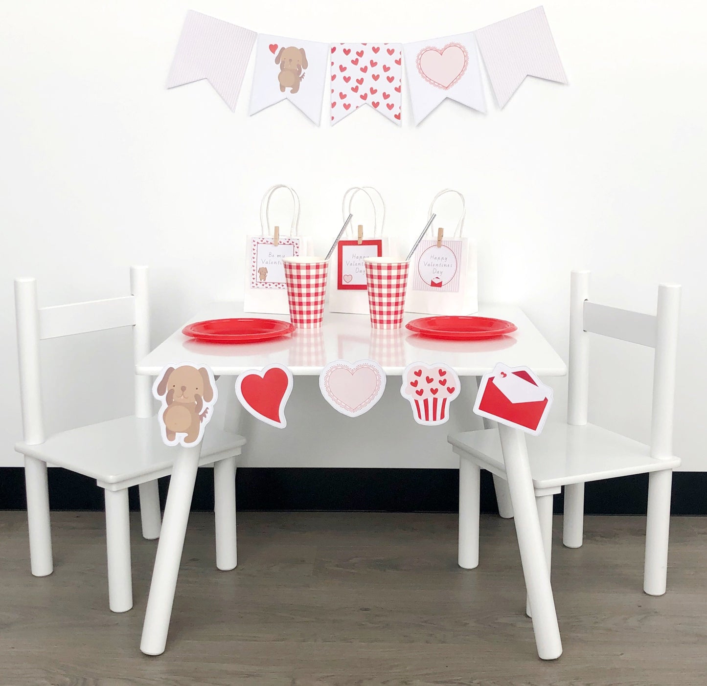 Valentines day celebrations set up with cute puppy and red love hearts accents, red plates and cups on white table. There is a matching bunting flag on the wall.