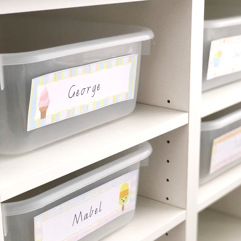 Clear plastic tubs have ice cream theme name plates on them and are stacked on white shelves in classroom.