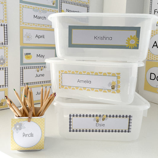 Classroom locker tubs stacked on white bench next to a jar with pencils. The items are labelled with yellow and grey themed labels. 
