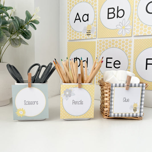 Stationery items including scissors, pencils, and a glue stick in labeled containers on a white surface with a decorative alphabet poster in the background.