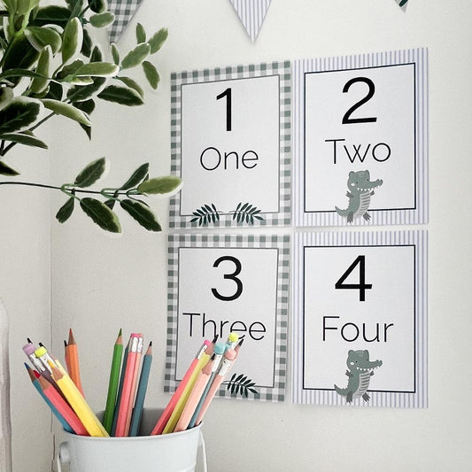 Crocodile theme educational number flash cards on white wall in classroom with pencils, bunting flag and plant on desk.