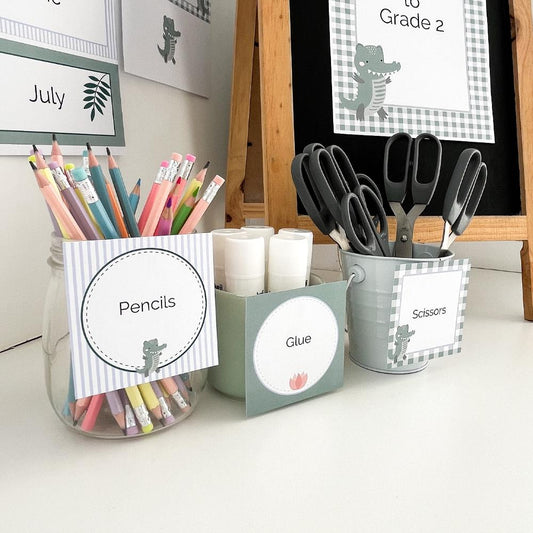 Square printable classroom labels in a crocodile theme on white bench with school supplies in caddies. 