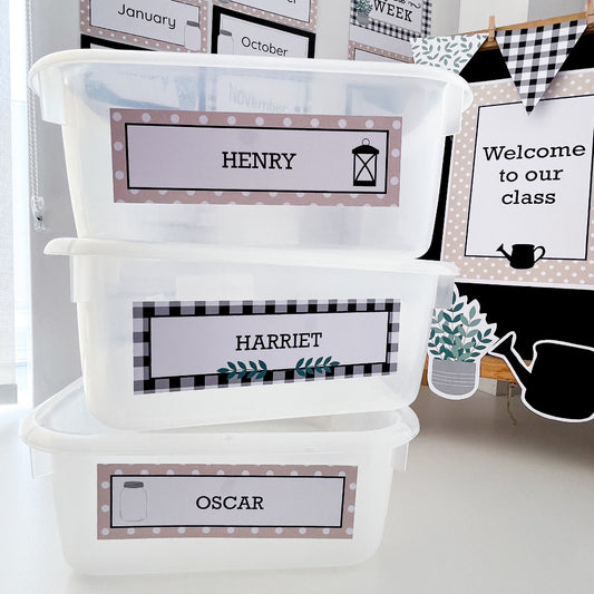 Three classroom tubs are stacked on a white shelf. They each have a black, white and beige name label on them. 