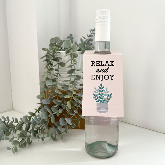 Wine label on a bottle of wine that says 'Relax and Enjoy'. The bottle is on a white bench with a pot plant behind it. 
