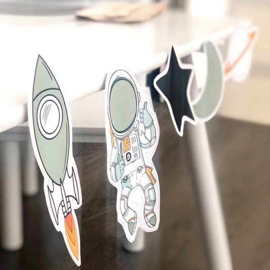 Space themed cut out shapes on white table. Shapes are rocket, astronaut, star, moon and planet. 