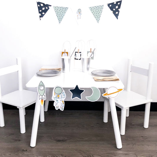 Space themed Party Decorations set up on white table with bunting flags and cut out shapes. 